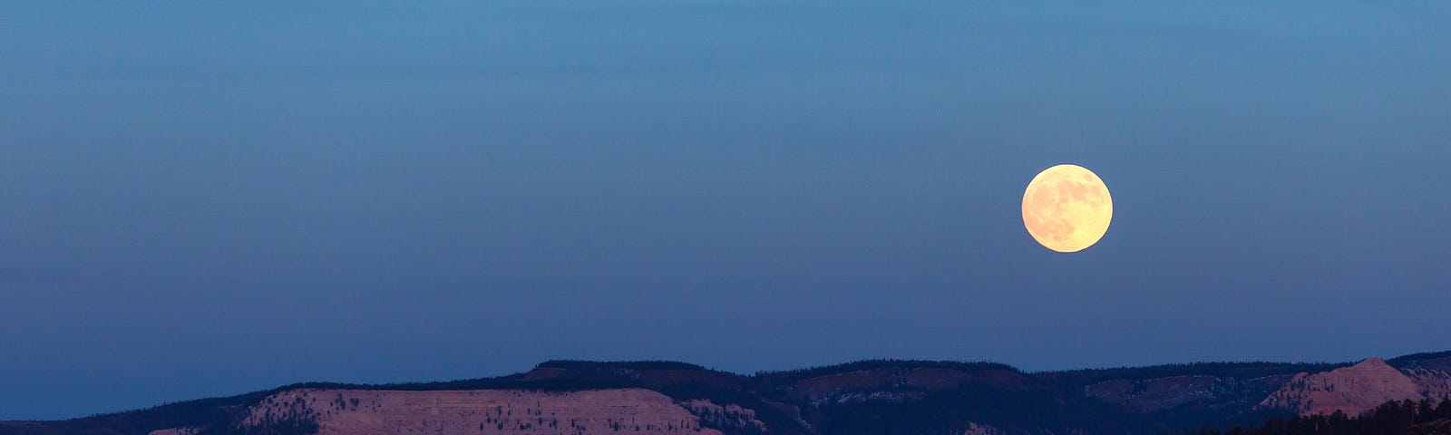 Moody photo of a full moon rising over a small mountain range at twilight.