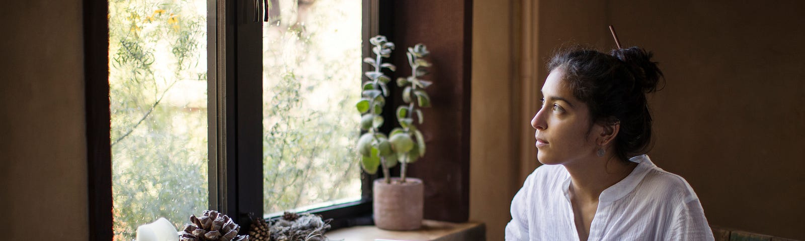 Young dark-haired woman looks out the window while on her laptop at a table, cup of tea in hand.