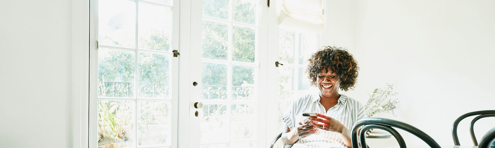 Smiling Black woman looking at her phone while sitting in a chair in her home.