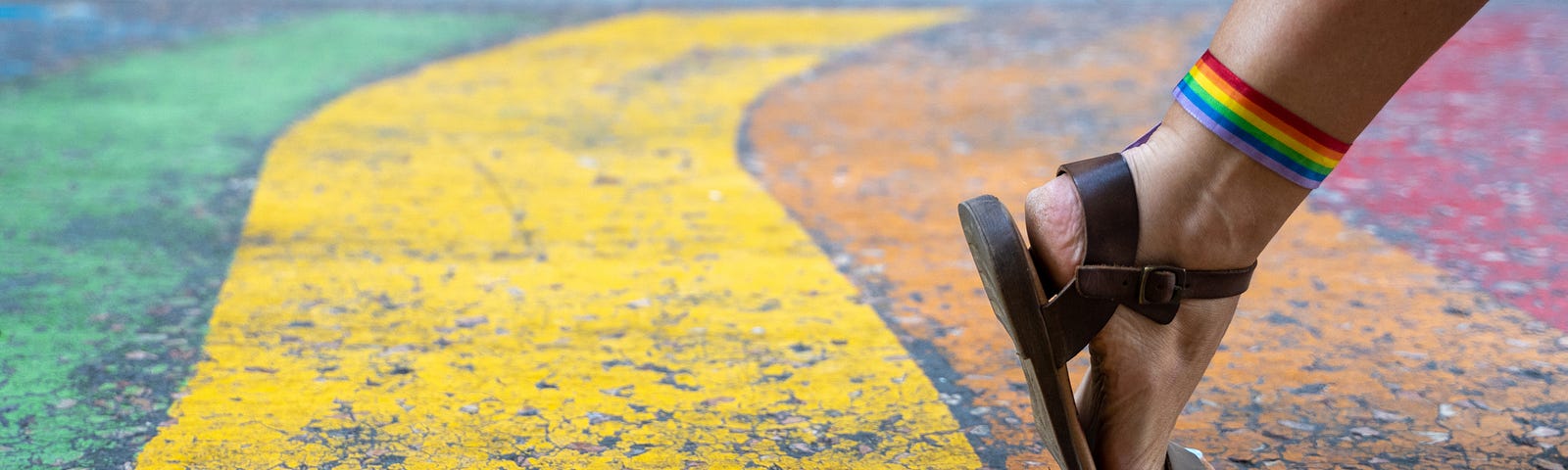 A woman’s foot with sandals and a rainbow flag tied to the knuckle making a step forward on a rainbow sidewalk.