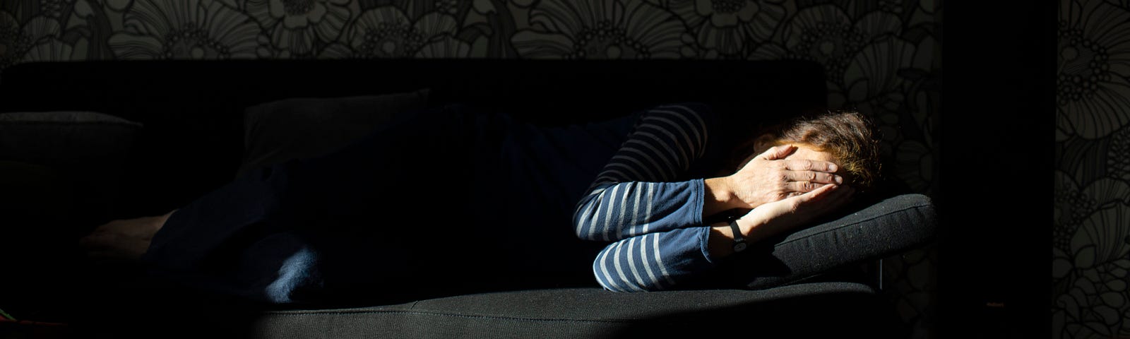 A woman lying sideways on a sofa covering her face with her hands.