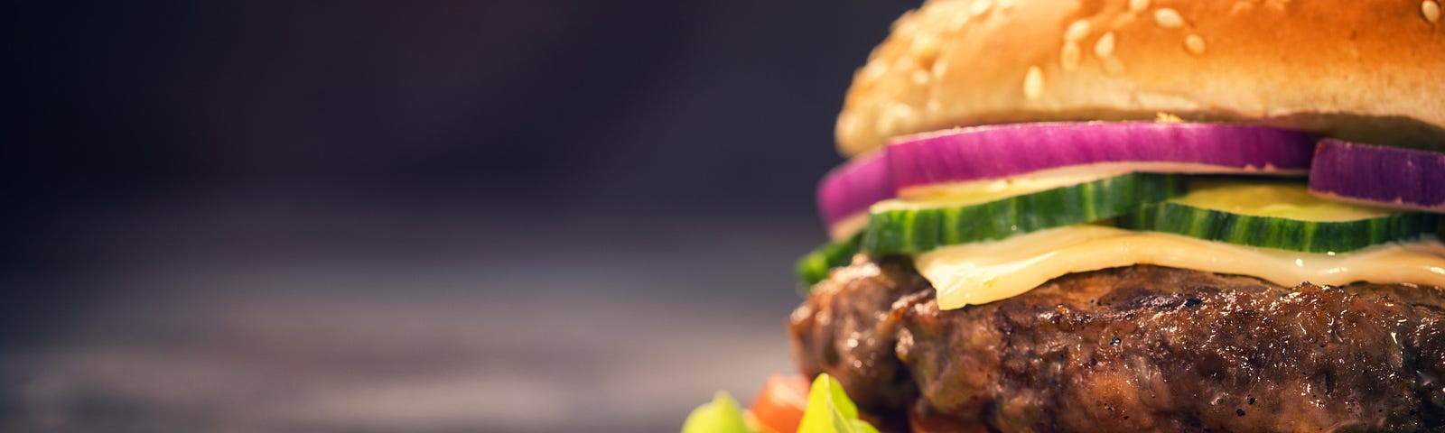 Close-up of a cheeseburger in a sesame seed bun with lettuce and sliced onions, tomatoes, and cucumbers on a black background