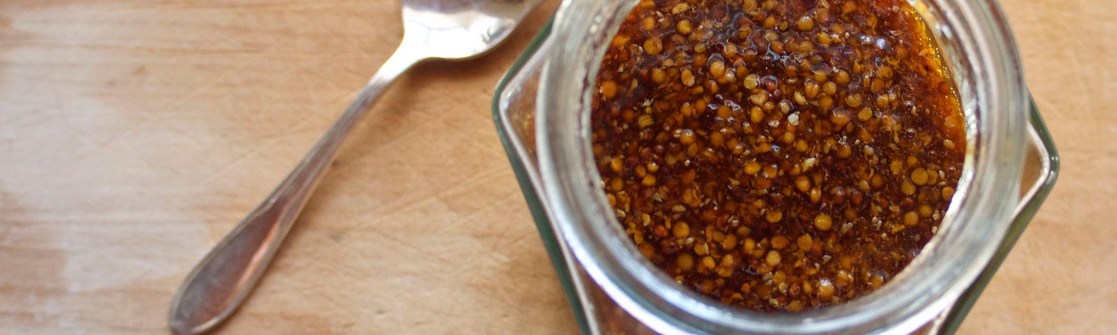 A photo taken from above of a dark, maple-y glass jar of mustard with visible mustard seeds and a small spoon resting nearby