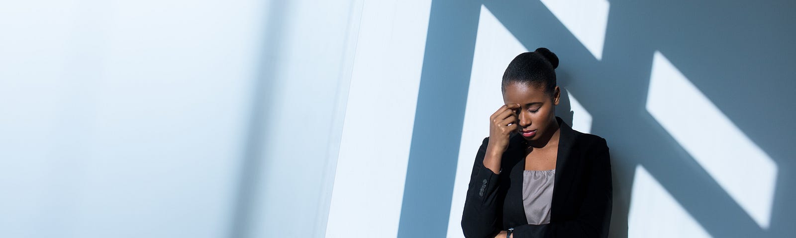 A photo of a stressed black woman at work. She is standing against a wall, wearing business casual clothes, eyes closed.