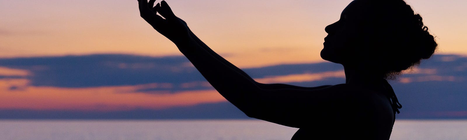 Silhouette of a woman with arms raised in front of her, palms up. She is standing in the foreground of a large body of water at sunset.