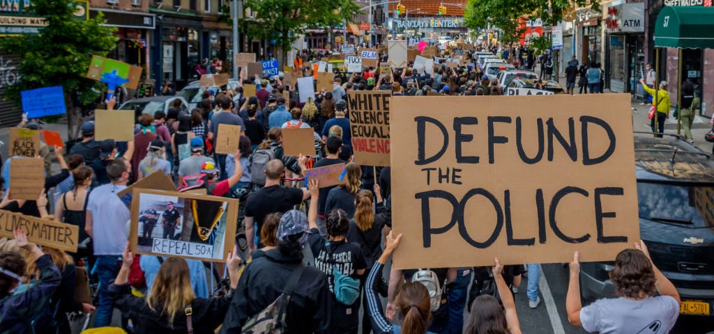 A photo of a protest. One big sign reads “DEFUND THE POLICE.”