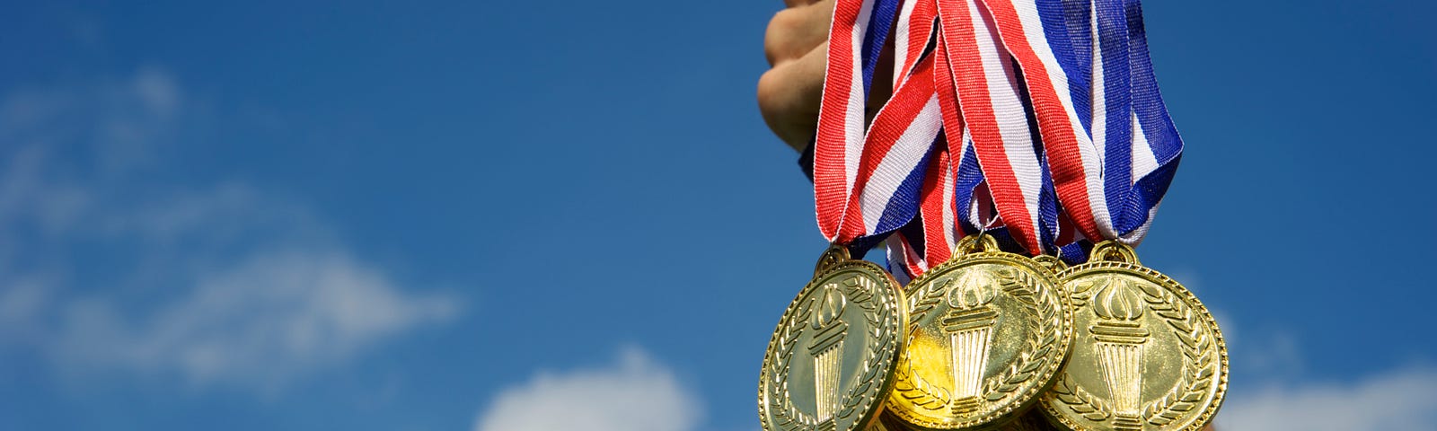 A hand holds up a bunch of gold, silver, and bronze medals against a bright blue sky.