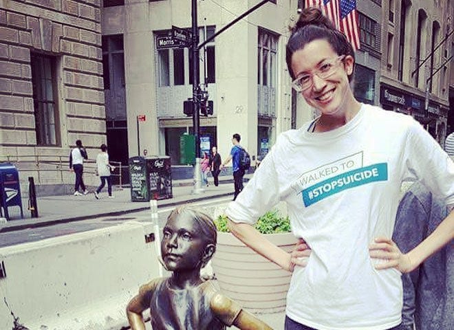 A young woman with glasses is smiling and standing next to the “Fearless Girl” statue in the Financial District of New York City. The woman matches the pose of the statue in that her arms are also on her hips and her legs apart.