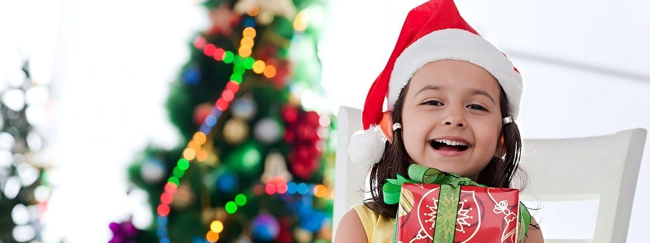 A little girl wearing a Santa hat sits in front of a Christmas tree with a present in her arms.