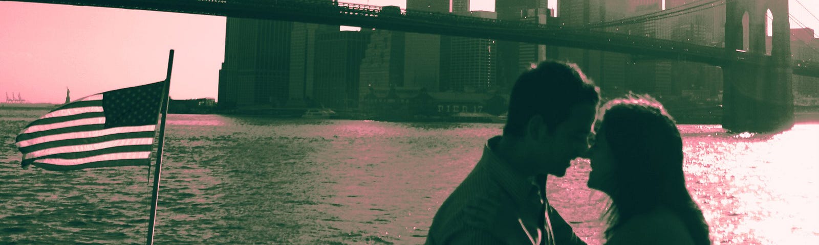 A photo of a couple standing by the water overlooking a bridge and a city.