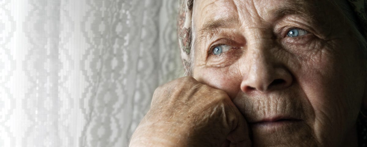 Portrait of a pensive elderly woman with her hair swept back from her face. She is next to a window covered by lace curtains. The woman looks sad. Dramatic lighting.