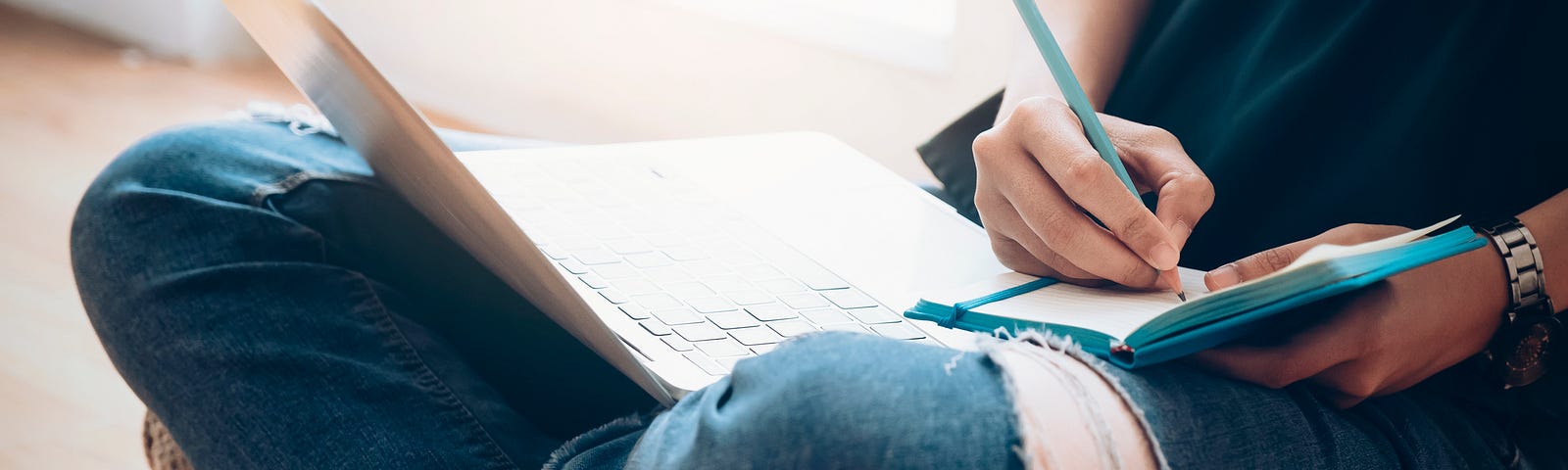 A photo of a woman writing in her journal while her laptop is on her lap.