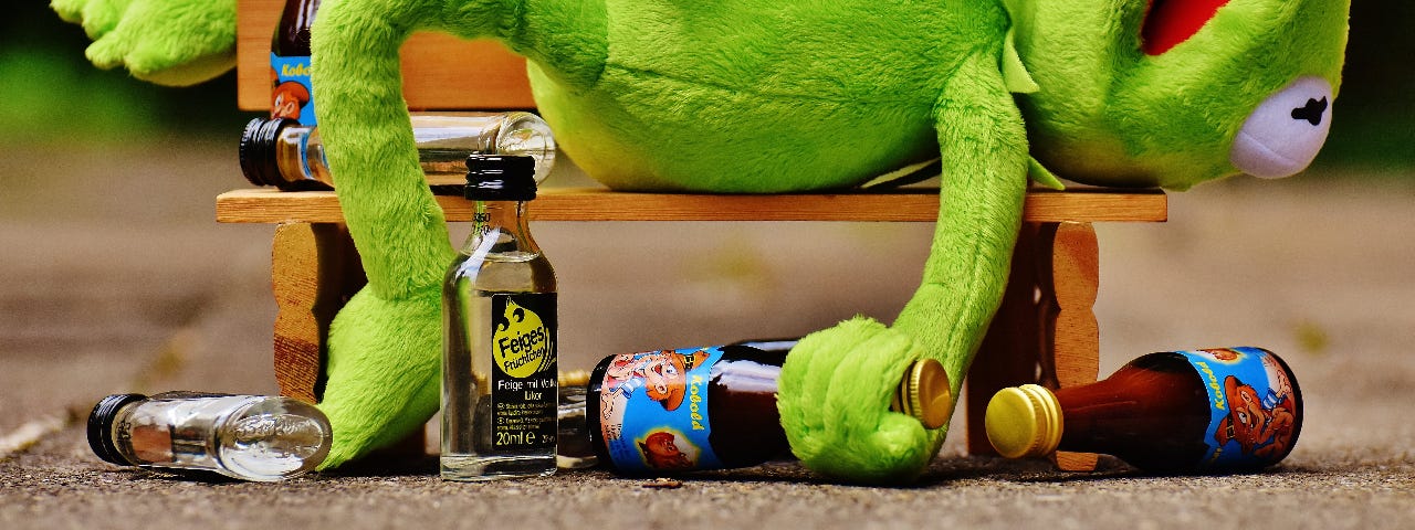 Kermit The Frog lying on wooden bench surrounded by glass bottles. Kermit appears drunk with a bottle of clear bottle presumed to be vodka in one hand and a brown bottle presumed to be beer in the other. He’s lying on his back with his mouth open and one leg hanging over the back of the bench.