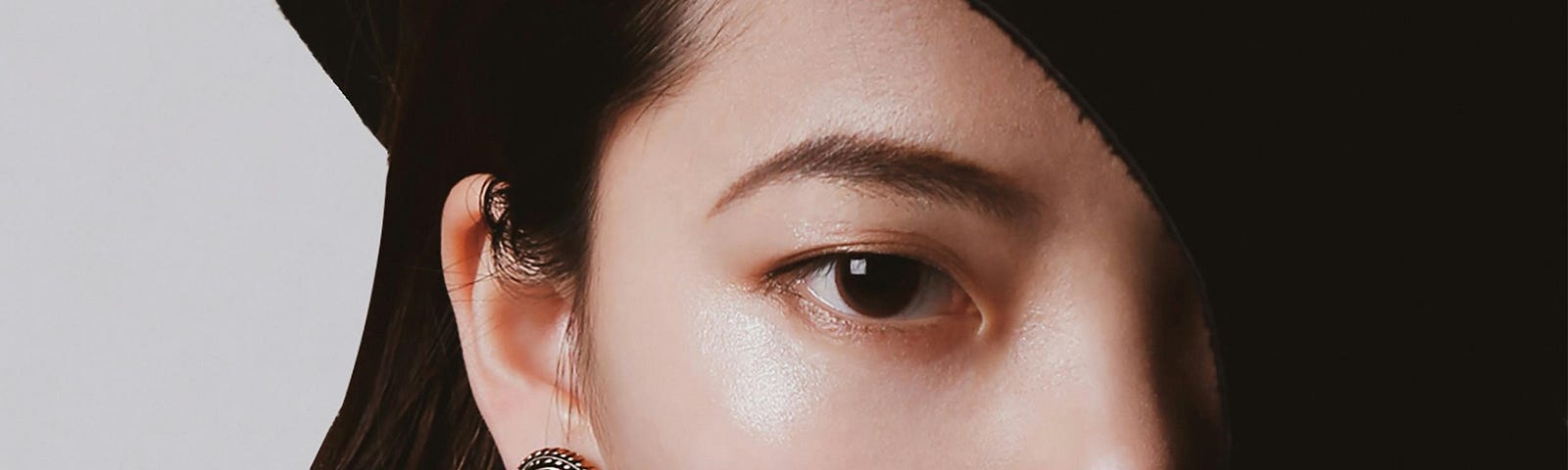 A woman of Asian descent with dark hair stares in a close-up shot. She’s wearing a black wide-brimmed hat, slanted down over her left eye, and her outfit is black.