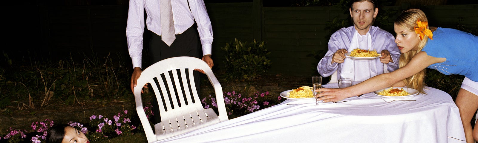 One woman falls off a chair and pulls the dining table cloth as her friends awkwardly try to help her.