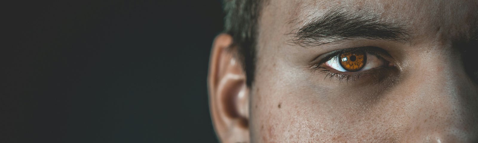 A close-up image of a man with reddish-brown eyes. Half of his face is visible and the other half is obscured. The backdrop is black.