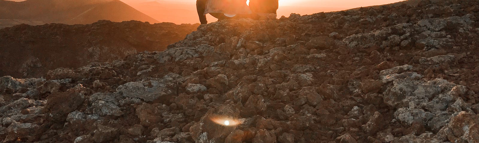 Sunset or sunrise over a rocky environment with mountains in the distance. Two people, probably male and female, sitting near each other in silhouette against the bright sun.