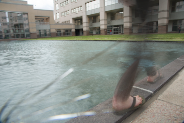Blurry image of feet walking along a pool of water with a building in the background.