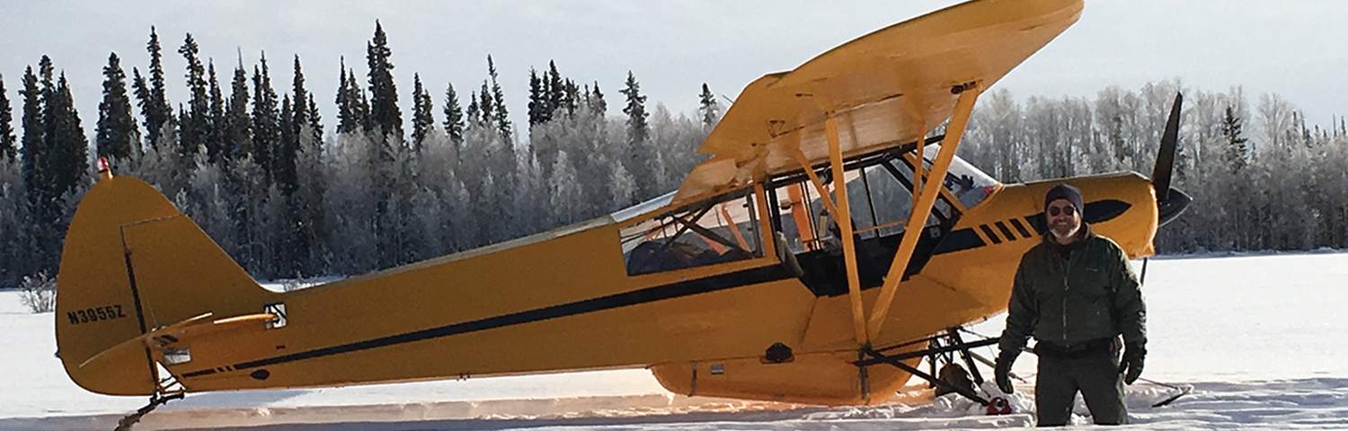 Photo of a yellow airplane on skis in the snow with the pilot.