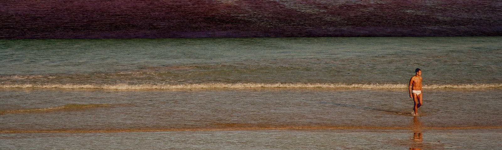 A photo of a man walking on the beach. The sky is dark with red clouds.