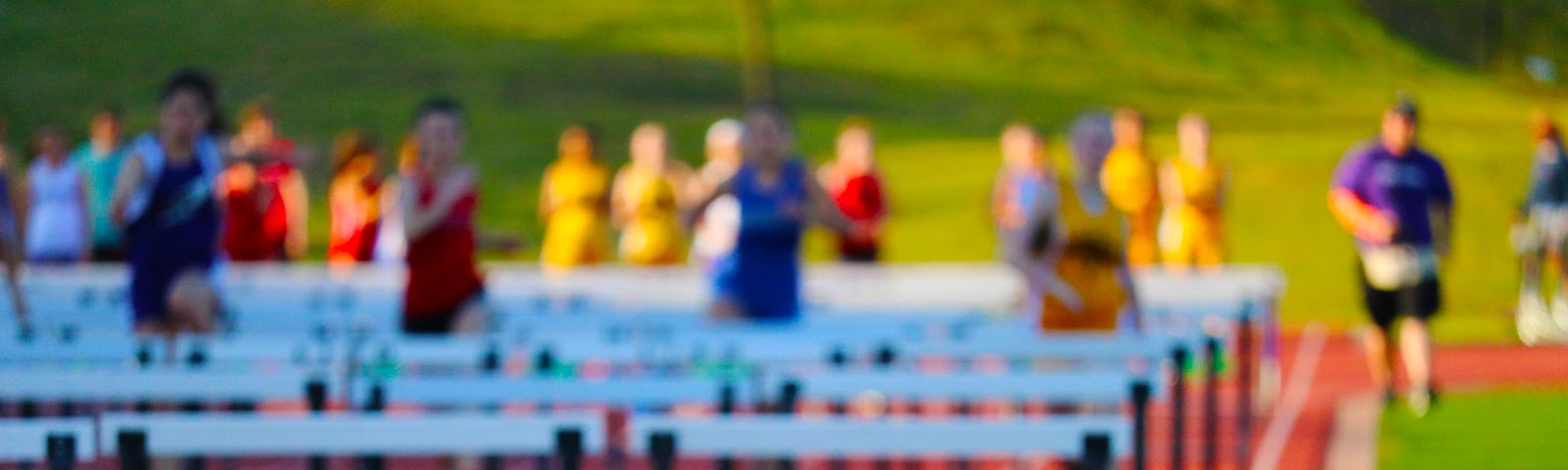 Adolescent children in colorful pennies run on a track with hurdles. The hurdles are in the foreground on a red/brown track and the kids are positioned toward the back of the photo, out of focus. They were pennies that are blue, yellow, red, and purple, and they appear to be running toward the camera.