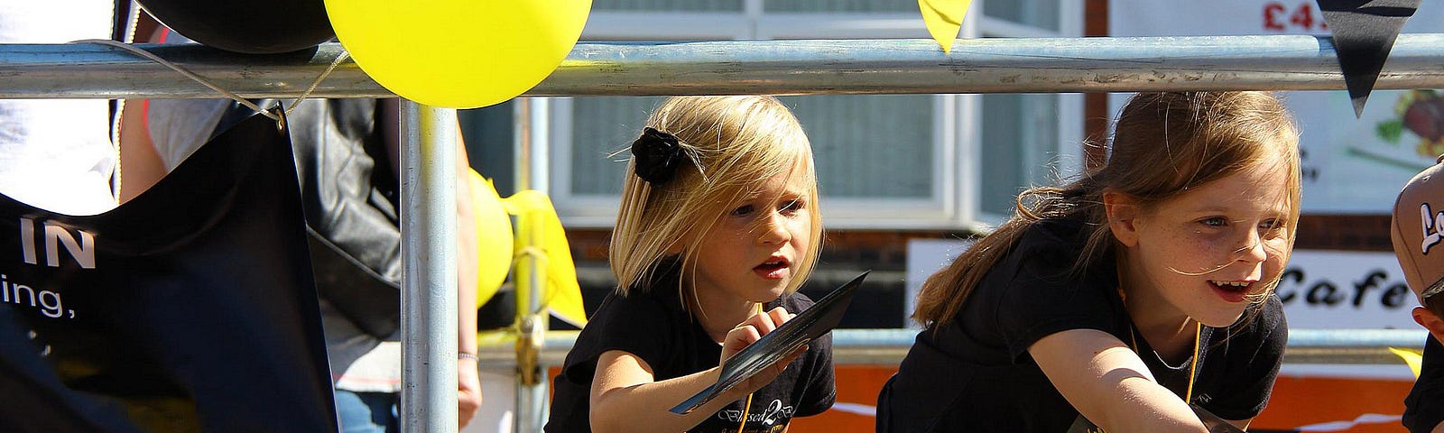 Two pre-teen girls handing out leaflets on a nice summer day. They are both bend down to pass the leaflet from between two horizontal bars in the fence. It is impossible to see what is on the leaflet. Yellow and black flags and balloons can be seen in the periphery of the picture.