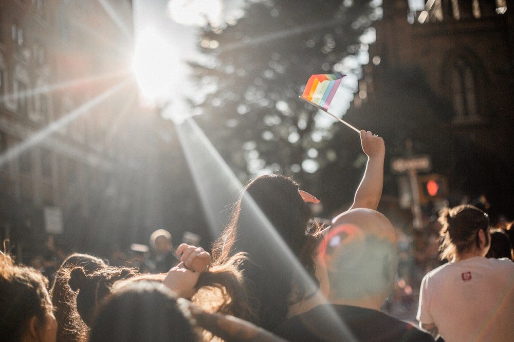 A small rainbow flag is raised above a crowd in the sunlight.