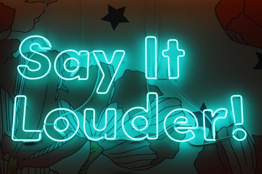 The sentence “Say It Louder!” in large, green neon lights.