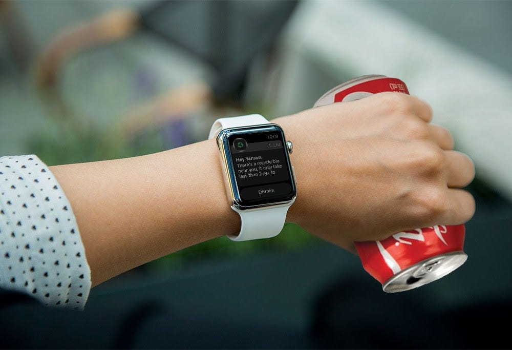 A woman’s hand holding a soda can and wearing a smart watch. The screen of the watch says “There is a recycling can near you. It only takes less than 2 seconds to…”