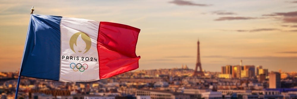 A French flag branding the 2024 Olympics’ logo flies in the forefront of an out-of-focus photo of Paris and the Eiffel Tower.