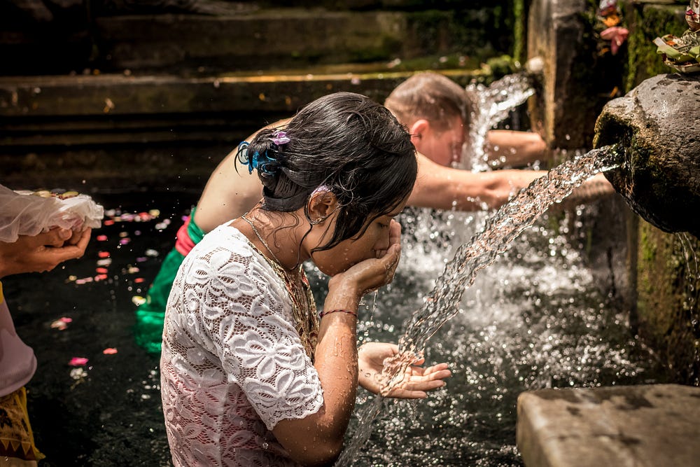 A young man and woman stand in an outside fountain pool while drinking water from it’s spout with cupped hands.