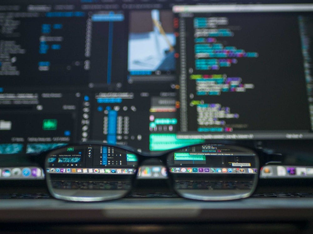 Ao fundo monitores com tela preta e código fonte nas cores brancas, azul, verde e vermelho, na frente um óculos direcionado para telas.