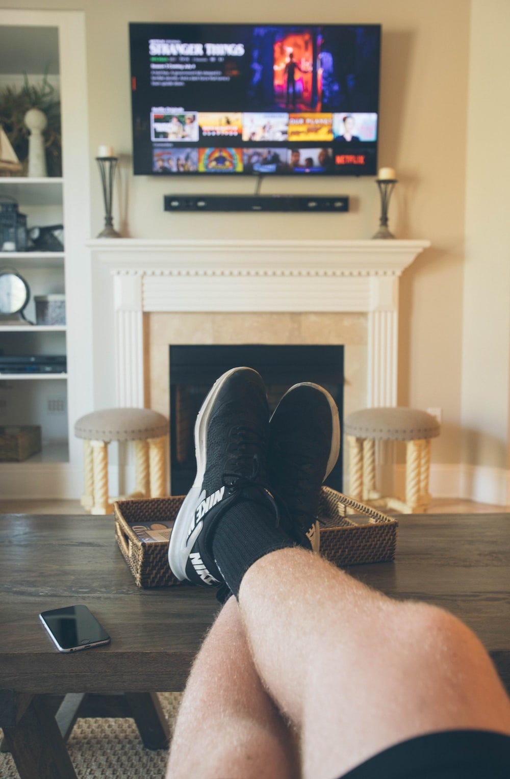 First-person POV: A man watching TV, pair of feet and a phone on the table.