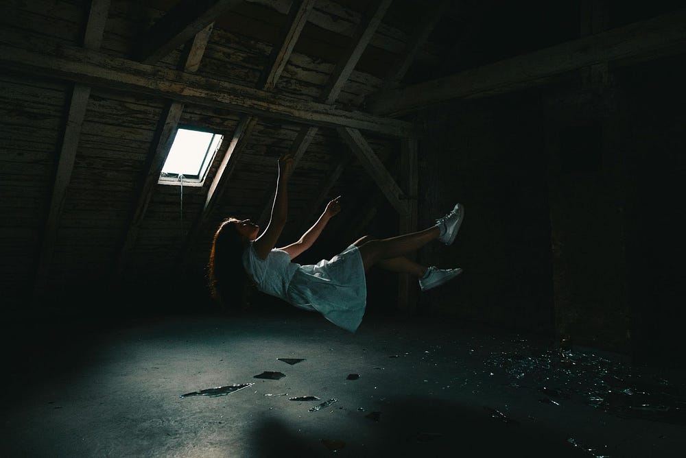 Picture of a girl in a blue dress and white shoes falling in a dark attic. Her arms are reaching upwards, hair is falling downwards. The attic has one small window on the left side and has a concret floor that has debris on it.