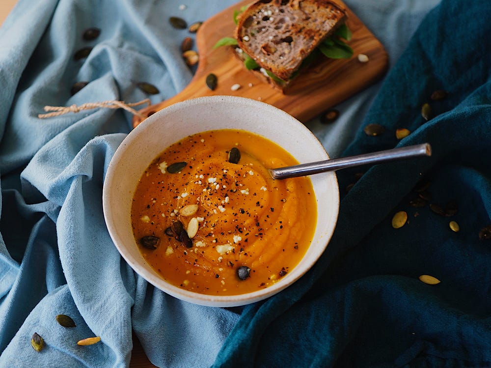 A white bowl filled with a thick creamy pumpkin colored soup sits on a blue cloth next to a single slice of bread on a miniature breadboard.