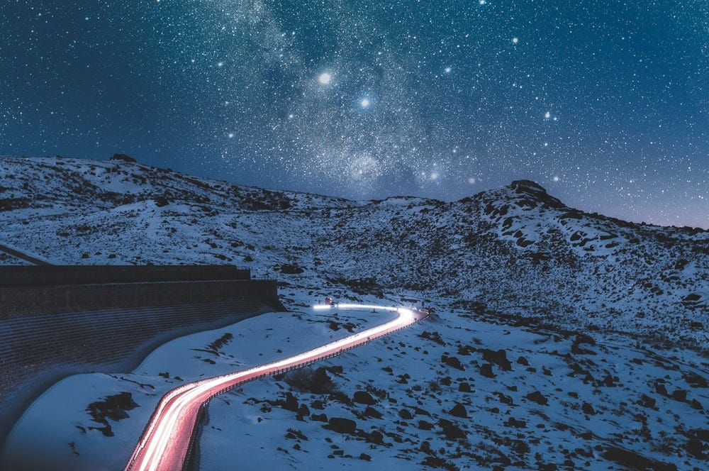 A windy road where there are mountains with snow ahead and stars in milky ways above. Gorgeous!