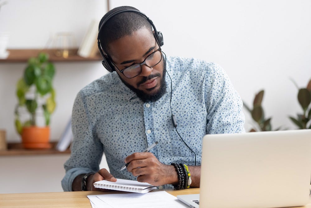 Homem negro com fone de ouvidos, olhando para seu laptop e tomando notas usando um bloco de papel e caneta, como se estivesse assistindo a alguma palestra ou curso no computador.