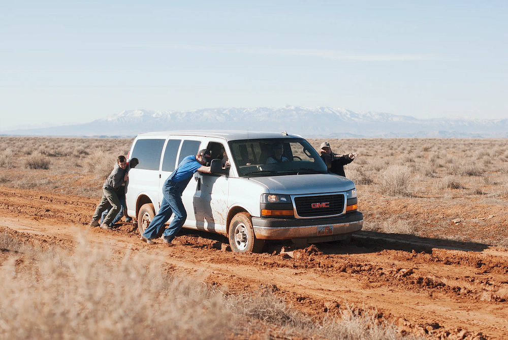 four people pushing a car on a dirt road
