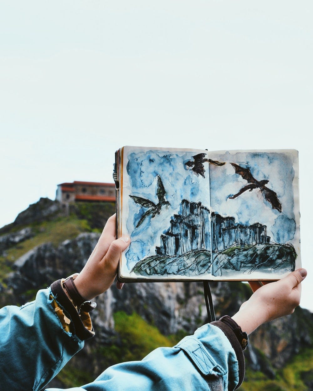 Image of a person’s arms and hands holding open a sketch book drawing of dragons soaring above a craggy cliff. In the background is an actual cliff with a low building at the top.