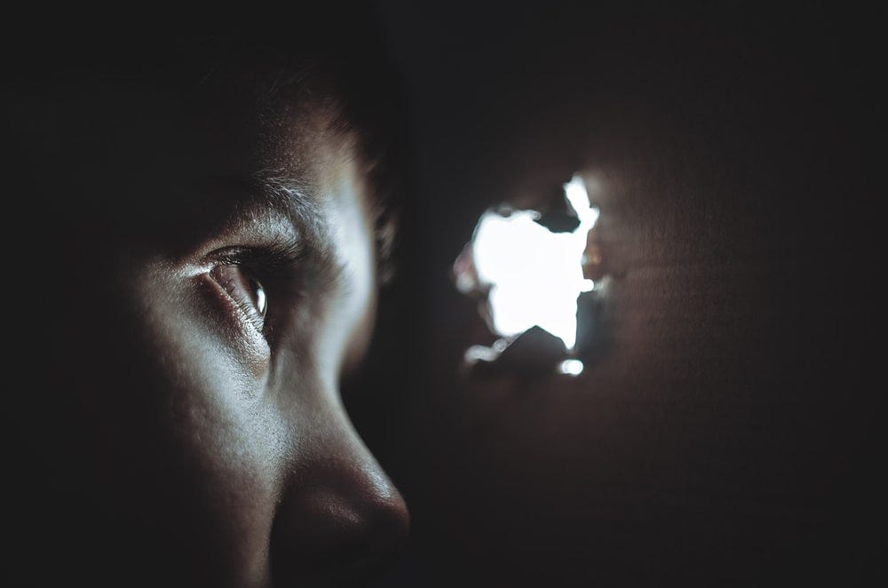 Image of a person in darkness looking through a small hole toward the light.