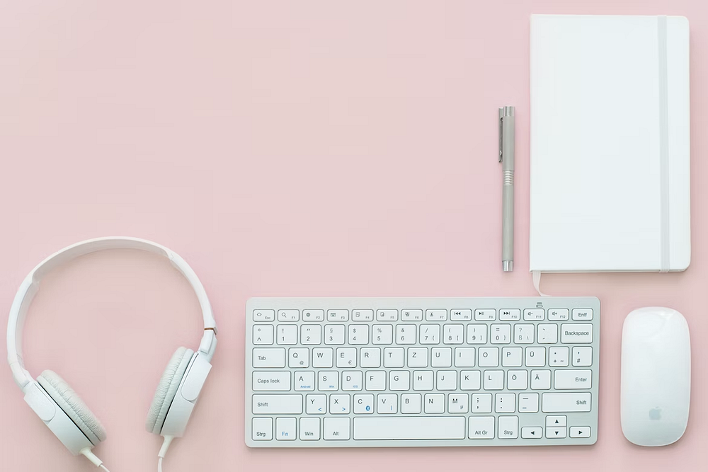 Photo of a white pair of headphones, keyboard, pen, notebook, and computer mouse against a light pink background.