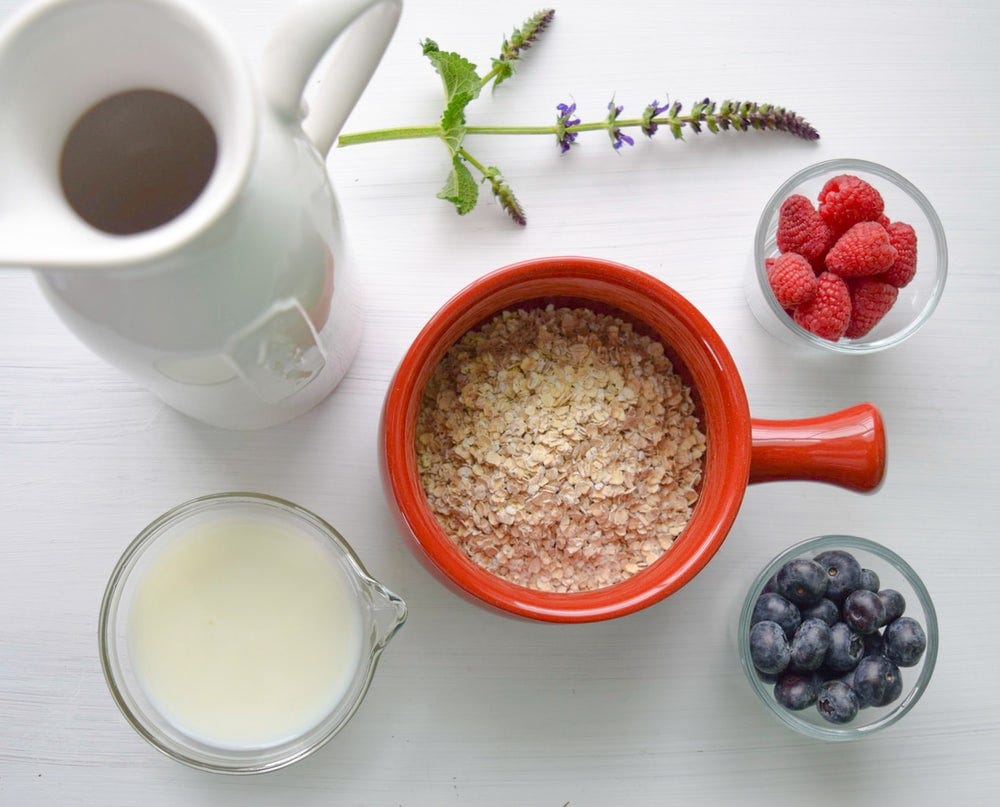 Upper view of a flask, a glass of milk, a bowl of oats and 2 small bowls of berries on a white table.