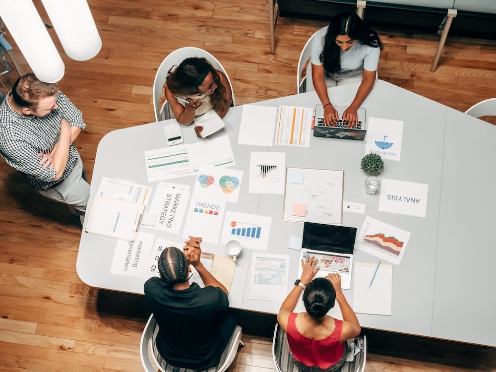 Cinco pessoas, em uma sala de reunião, ao redor de uma mesa, analisando informações em notebooks e vários papeis com gráficos e dados de pesquisas.