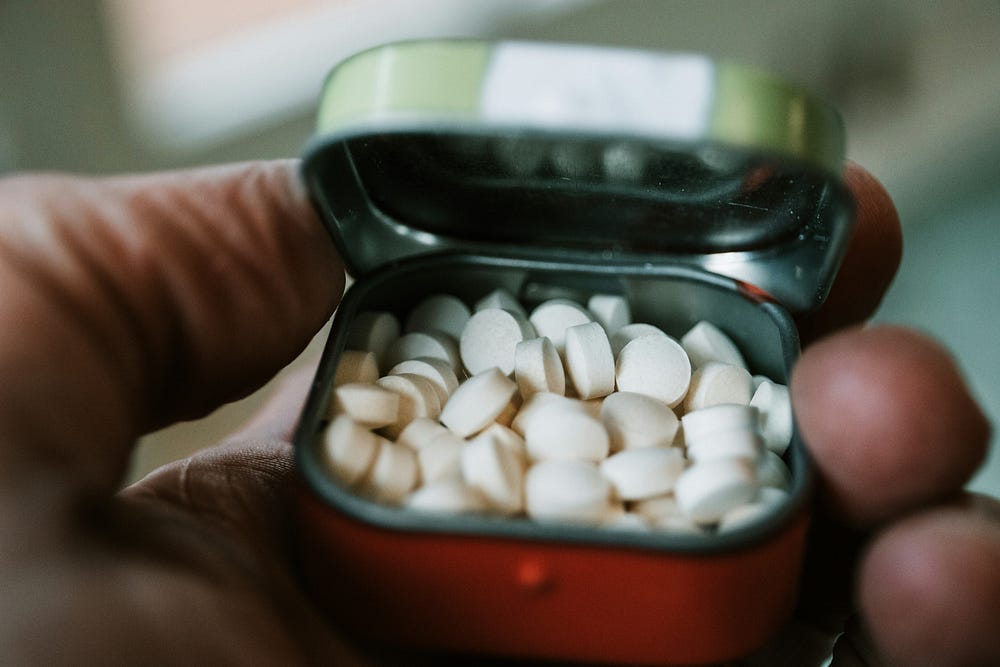 A hand holding an open red pill box with a silver metal top containing  white tablets.