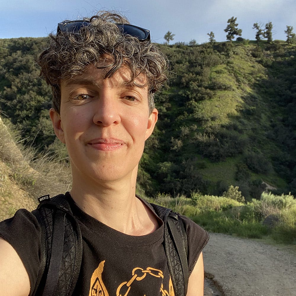 Portrait of Emily, smiling with messy curls and sunlit eyes, on a green mountainside in Southern California.