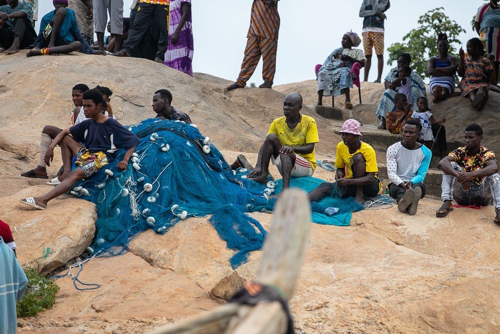 Several fishers sit on a sandy shore with a large blue fishing net.