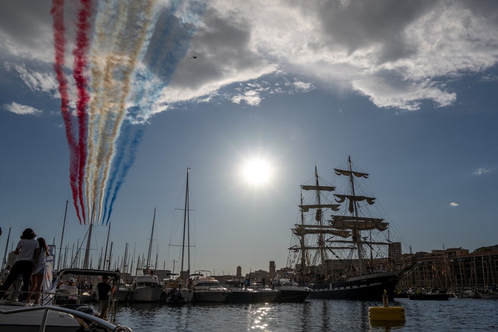 The Belem ship enters the Old Port to bring the Olympic flame to Marseille for Paris 2024 olympic games. Streaks of red, white, and blue smoke can be seen in the sky.