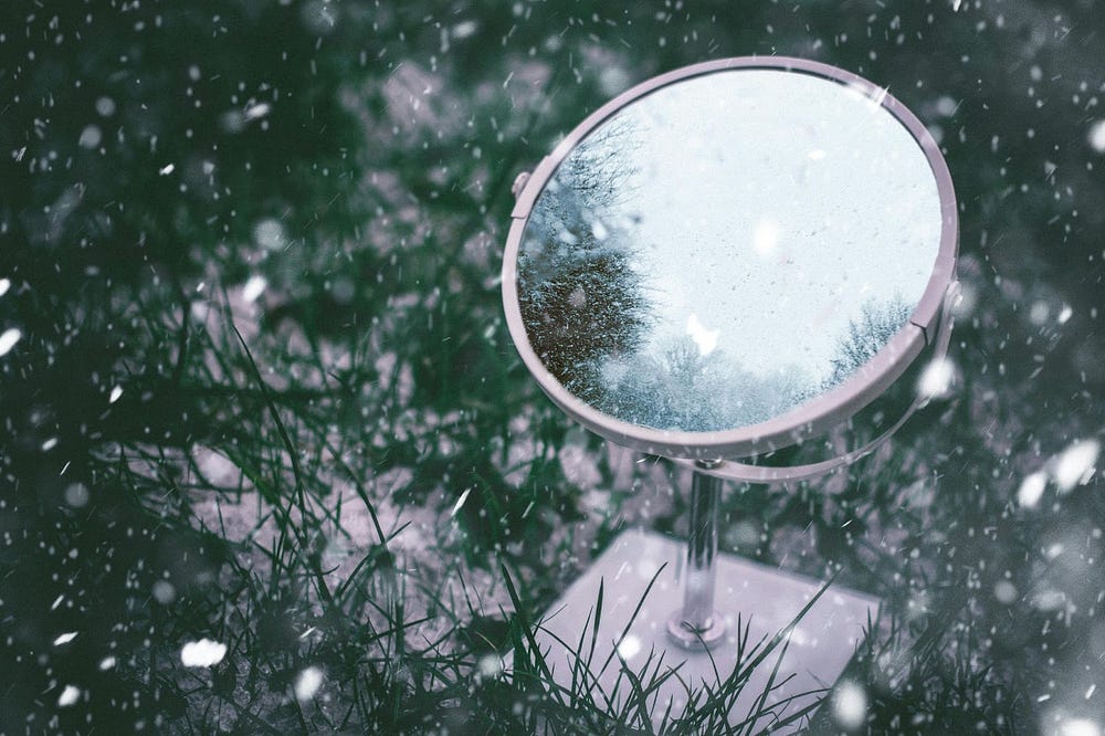 Photo of a round makeup mirror in dark green grass which has been lightly covered in snow. Snow is falling on to the mirror and grass.