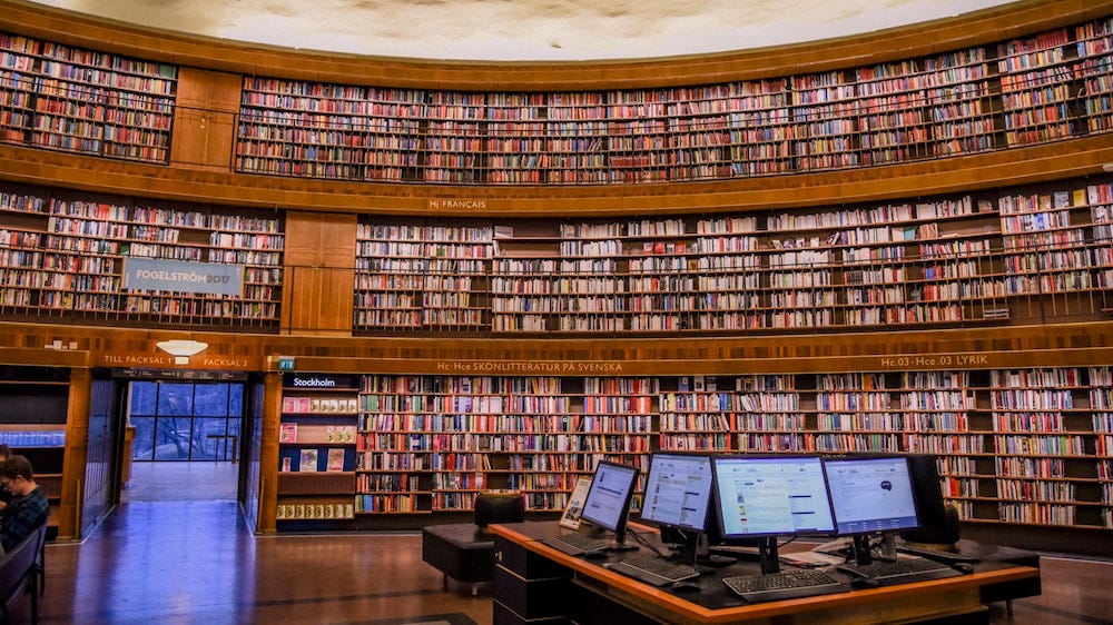 Foto de uma biblioteca com três andares de estantes de madeira, com livros, e uma mesa com diversos computadores para uso.