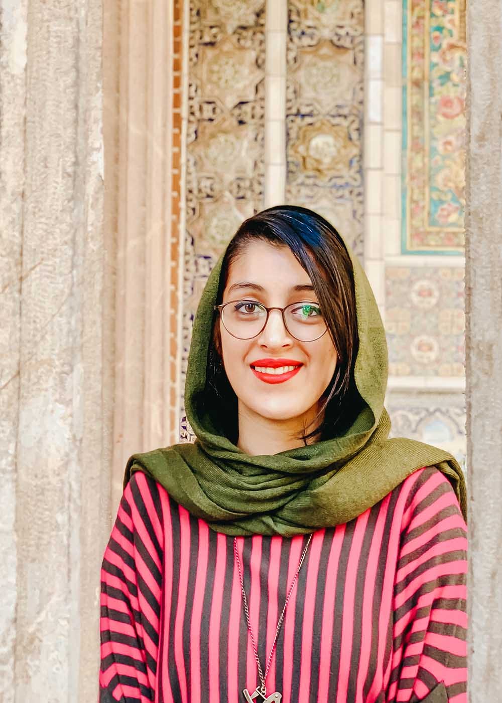 An Iranian girl with a scarf smiling in front of a historical wall.